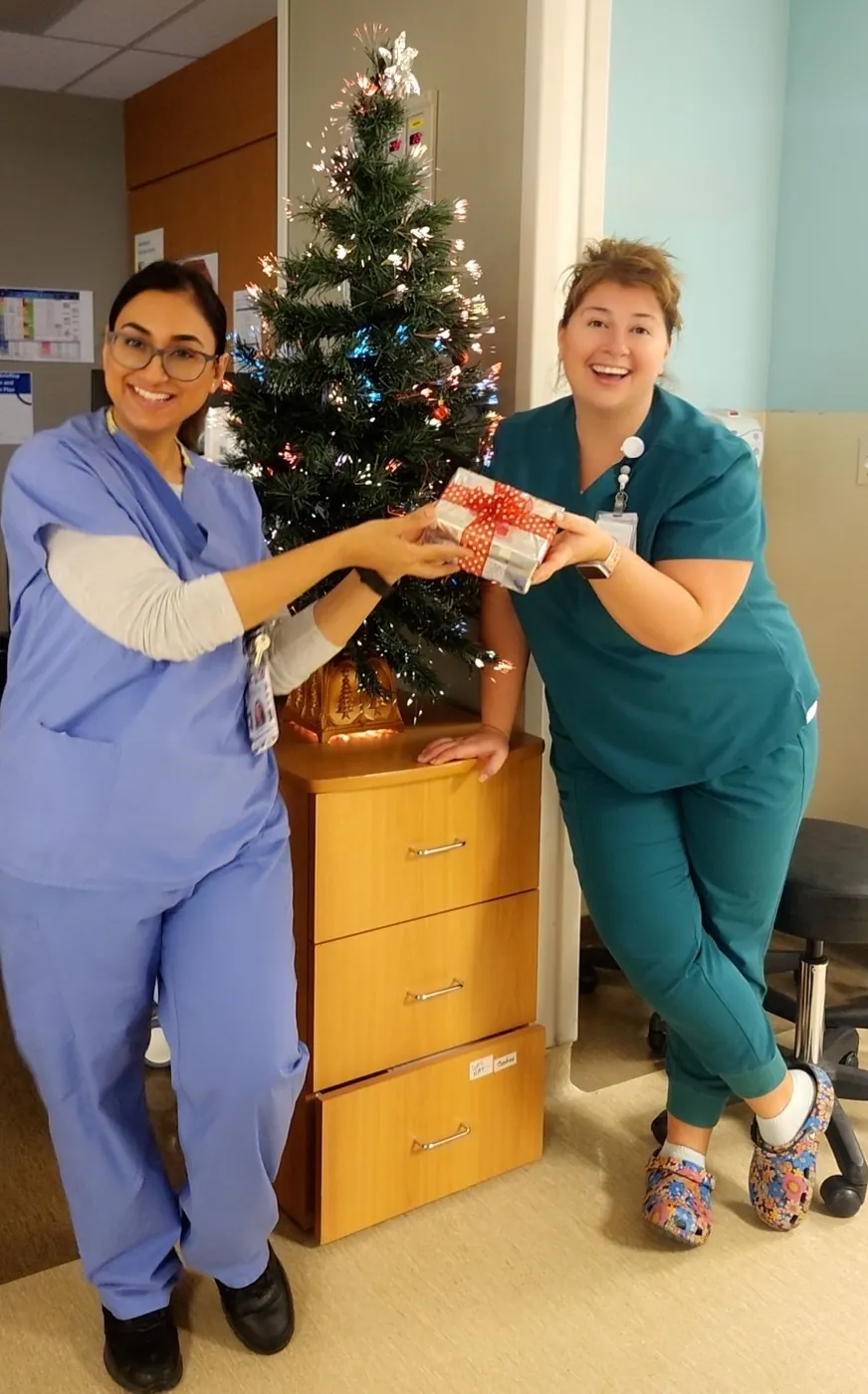 Jasraj and a colleague in scrubs exchanging a gift beside a small holiday tree