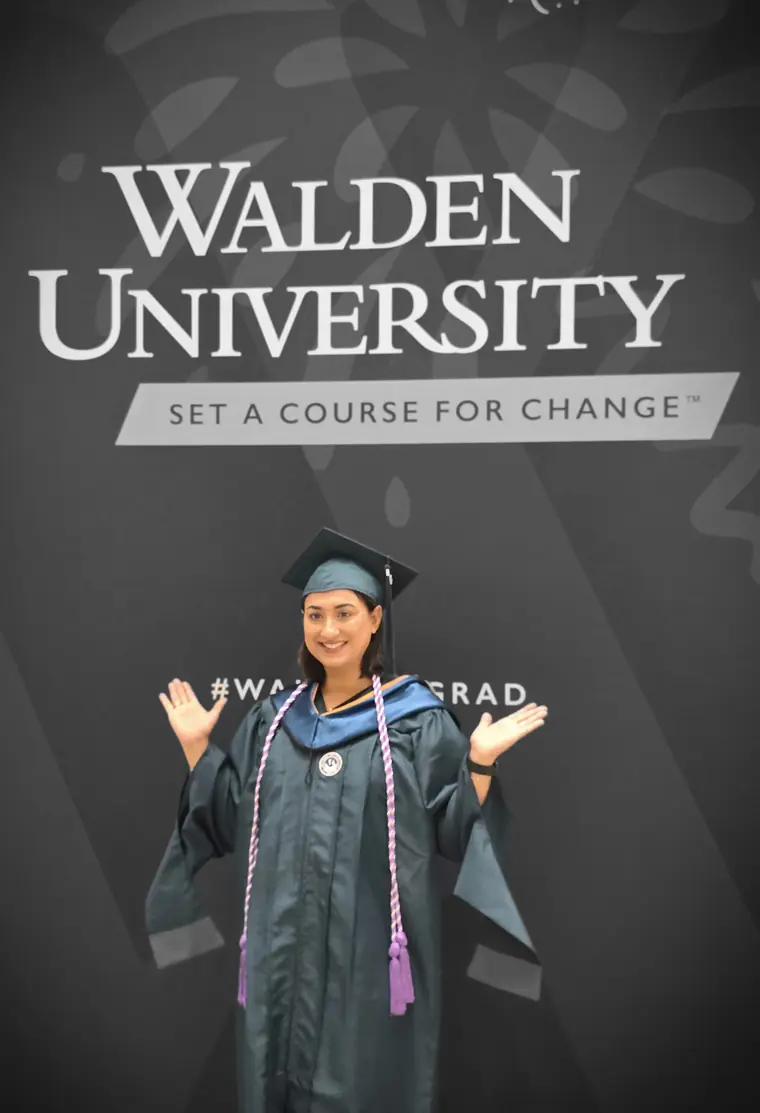 Jasraj in her graduation cap and gown celebrating in front of the Walden University backdrop